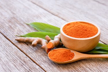 Tumeric powder, a food with curcumin,  in a wooden dish next to turmeric root on a wooden table.