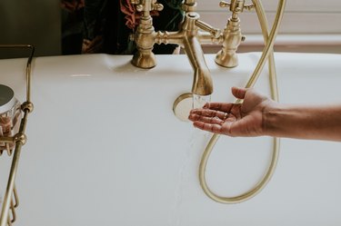 A woman checks the temperature of bath water by holding her hand under the faucet as the water runs