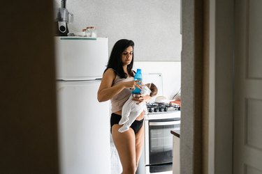 a person wearing black briefs and a tan tank top with long dark hair holds a newborn baby and a blue water bottle in front of a white fridge in a kitchen