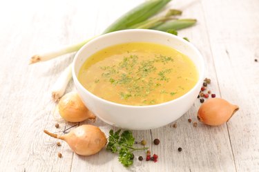 healthy vegetable broth made with fresh vegetables in a white bowl on wooden tabletop next to onions and fresh herbs