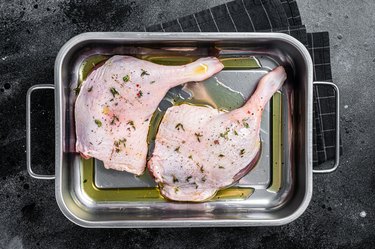 Raw meat, duck legs thighs with herbs and ingredients in a steel tray. Black background. Top view