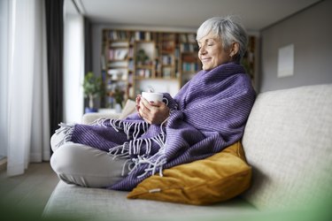 Older adult sitting on the couch wrapped in a purple blanket with a cup of tea because she feels colder as she gets older