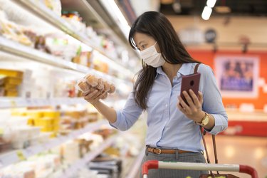 A woman in the grocery store wears a face mask as she examines a carton of brown eggs