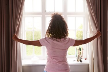 Rear View Of Woman Wearing Pajamas Opening Bedroom Curtains At Start Of New Day