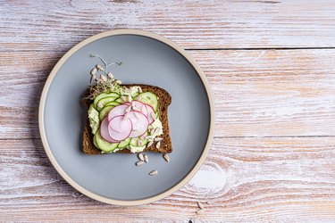 a slice of rye bread with avocado hummus dip and fresh cucumber, radish, sunflower seeds, and sprouts on a blue plate on a wooden table