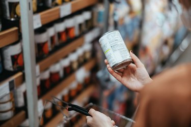 Over the shoulder view of young Asian woman carrying a shopping basket, grocery shopping in supermarket. Holding a tin can and reading the nutritional label at the back