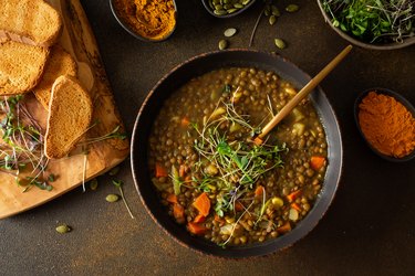 A wide shot of lentil soup in a dark brown bowl next to herbs and toasted bread