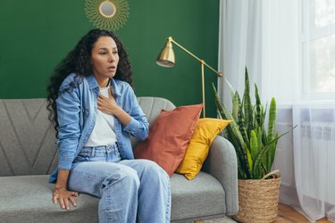 a person with long curly brown hair wearing jeans and blue jacket and sitting on a couch with their hand on their chest because they have shortness of breath from anxiety