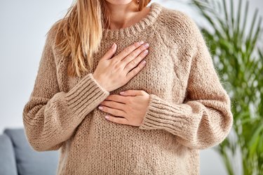 close view of a person with long blonde hair wearing a beige sweater and holding their hand over their heart because they have a flutter in their chest