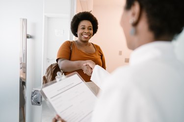 A smiling person wearing a dark orange t-shirt with a brown afro arriving at the doctor's office and shaking hands with the doctor.