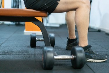 closeup of a man sitting on the edge of a workout bench with two dumbbells on the floor
