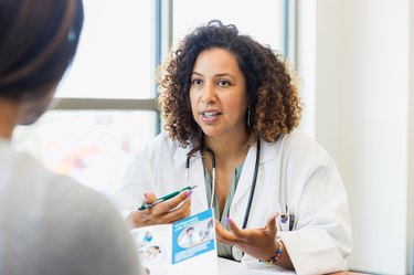 a doctor wearing a white lab coat and stethoscope talks to a patient in front of a window