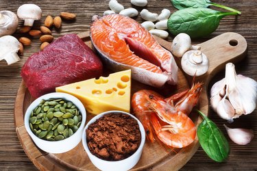 a wooden cutting board with foods high in zinc including salmon, cheese and shrimp, surrounded by garlic, basil leaves, mushrooms and nuts