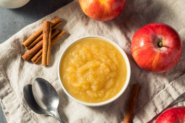 small white bowl of applesauce on a gray kitchen towel with apples and cinnamon sticks