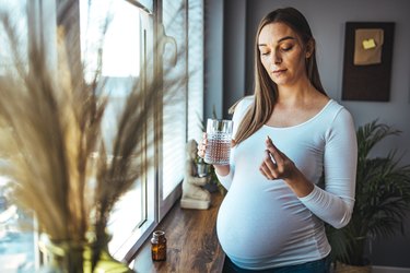 Pregnant person standing at window holding a glass of water and considering taking a pill, wondering about lishou coffee side effects and if the abortion pill makes you gain weight