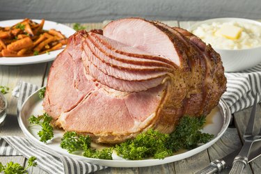 a close up of a Homemade Glazed Ham sliced on a white plate on a table next to a bowl of mashed potatoes and carrots