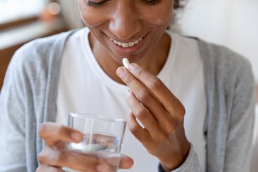 Close up photo of a smiling person holding a white probiotic and a glass of water wearing a white t-shirt and gray cardigan