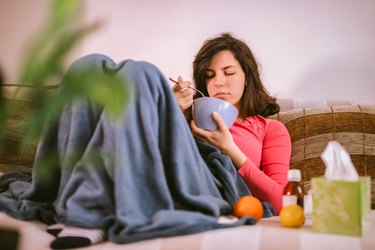 a young adult with the flu sits on the couch under a blanket eating a bowl of soup