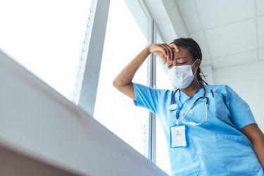 A doctor wearing blue scrubs and a face mask looks stressed in a hospital setting holding their hand to their forehead