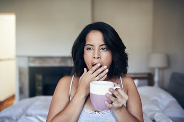 A woman drinking coffee and yawning as a symptom of too much caffeine