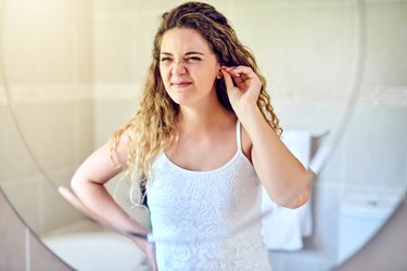 woman in her bathroom at home looking in the mirror and cleaning her ears with a cotton swab