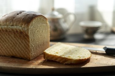 A close-up photo of a loaf of white bread with one slice cut off, on a wooden cutting board in the kitchen.