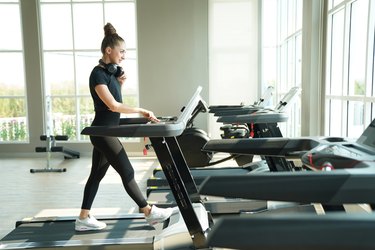 Young person with long hair in a bun and headphones walking on treadmill at gym