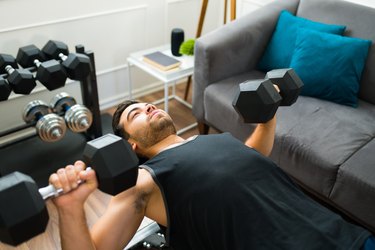 Close-up of person wearing black tank top doing a dumbbell bench press at home.