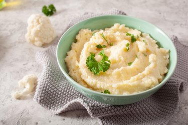 Cauliflower mash in a green bowl on gray countertop with fresh herbs prepared as an alternative to nightshade potato mash.
