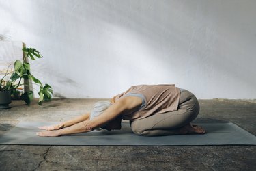 side view of a caucasian senior woman doing child's pose on a yoga mat in front of a white wall and plant