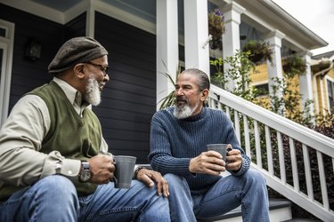 two older adults with beards drinking coffee while sitting on the front steps of a house