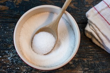 overhead photo of a wooden spoon in a white bowl of salt on a marble countertop next to a white dish towel with red stripes