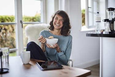 a woman with brown hair smiling and eating a bowl of oatmeal while sitting at her kitchen table