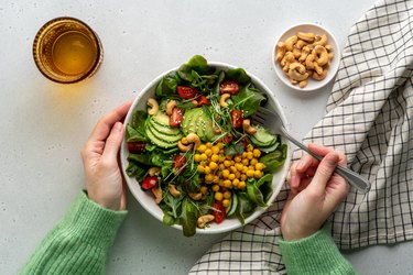 Top-down photo of a big bowl of green salad with cherry tomatoes, cucumber, avocado, chickpeas, cashews and white sesame seeds on top