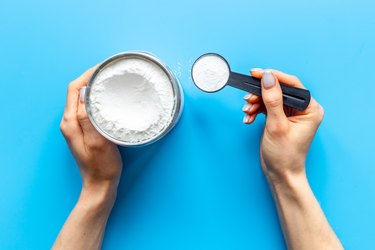 an overhead photo of hands holding a scoop with whey protein next to a jar of more powder on a bright blue background