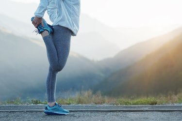 a close up of the lower body of an athlete wearing leggings, a running jacket and running sneakers doing a sartorius stretch outside with mountains in the background