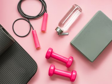 Yoga mat, weights, jump rope, water bottle and exercise journal on pink background.