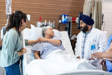 an older adult in a hospital bed holds a younger adults hand while advocating for themselves while talking to a doctor