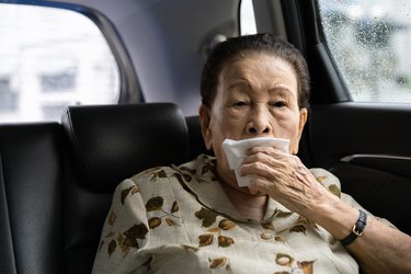 An older adult sitting in the backseat of a car and holding a napkin over their mouth because they have excessive saliva