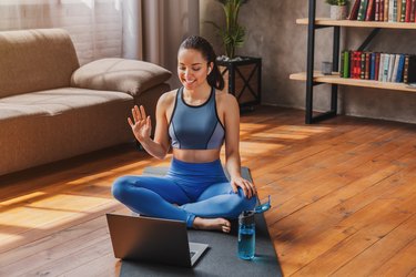 woman sitting down on a black yoga mat and waving at a laptop t in her living room