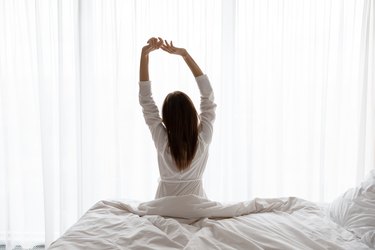 Beautiful woman sitting on bed, stretching arms in the morning after sleeping with lower back pain