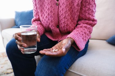close view of a person wearing a pink sweater holding a glass of water in one hand and two probiotic pills in the other hand