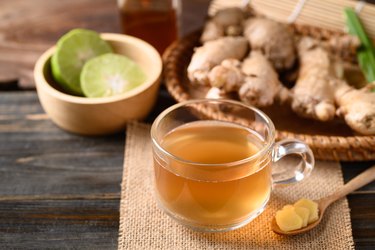 a glass cup of ginger tea with lime and honey and raw ginger in the background on a on wooden cutting board