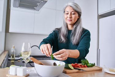 a person with long grey hair in the kitchen making a salad with vegetables