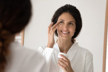 an adult wearing a white robe smiles in the mirror while applying eye cream