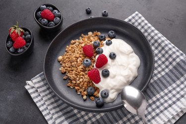 Yogurt with baked granola and berries in black ceramic plate on dark stone background