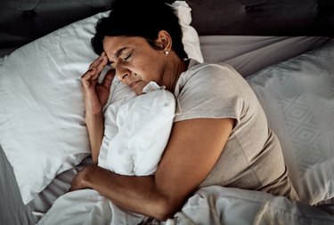 a middle-aged person with short brown hair and earrings wearing a tan t-shirt and lying in bed holding their head because they have a headache when lying down