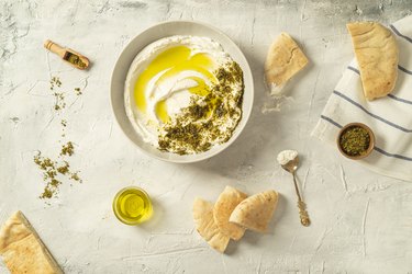 Popular middle eastern appetizer labneh or labaneh, soft white goat milk cheese with olive oil, hyssop or zaatar, served with pita bread on a grey table, top view,