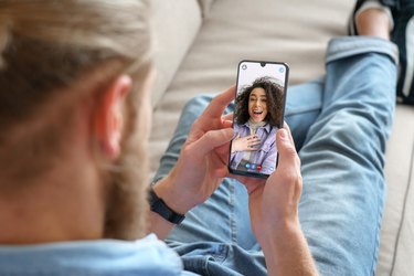 Young person sitting on sofa holding smartphone communicating with person on mobile screen, making video call.