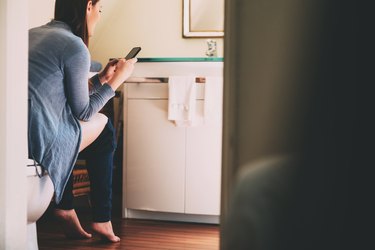 a person with long brown hair wearing a sweater and jeans and sitting on the toilet while texting